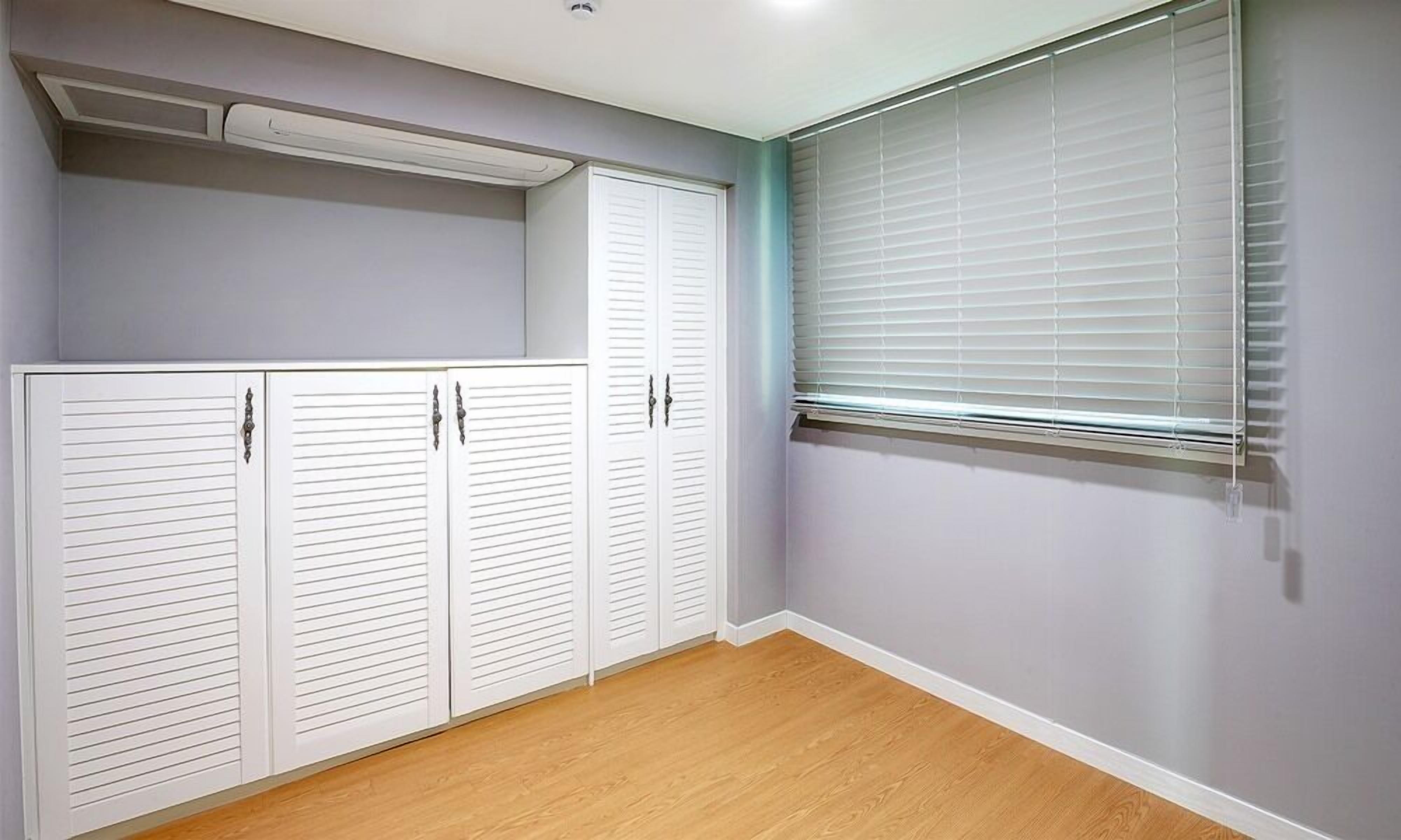 A room with a wooden floor, a window with blinds, and white louvered closets.