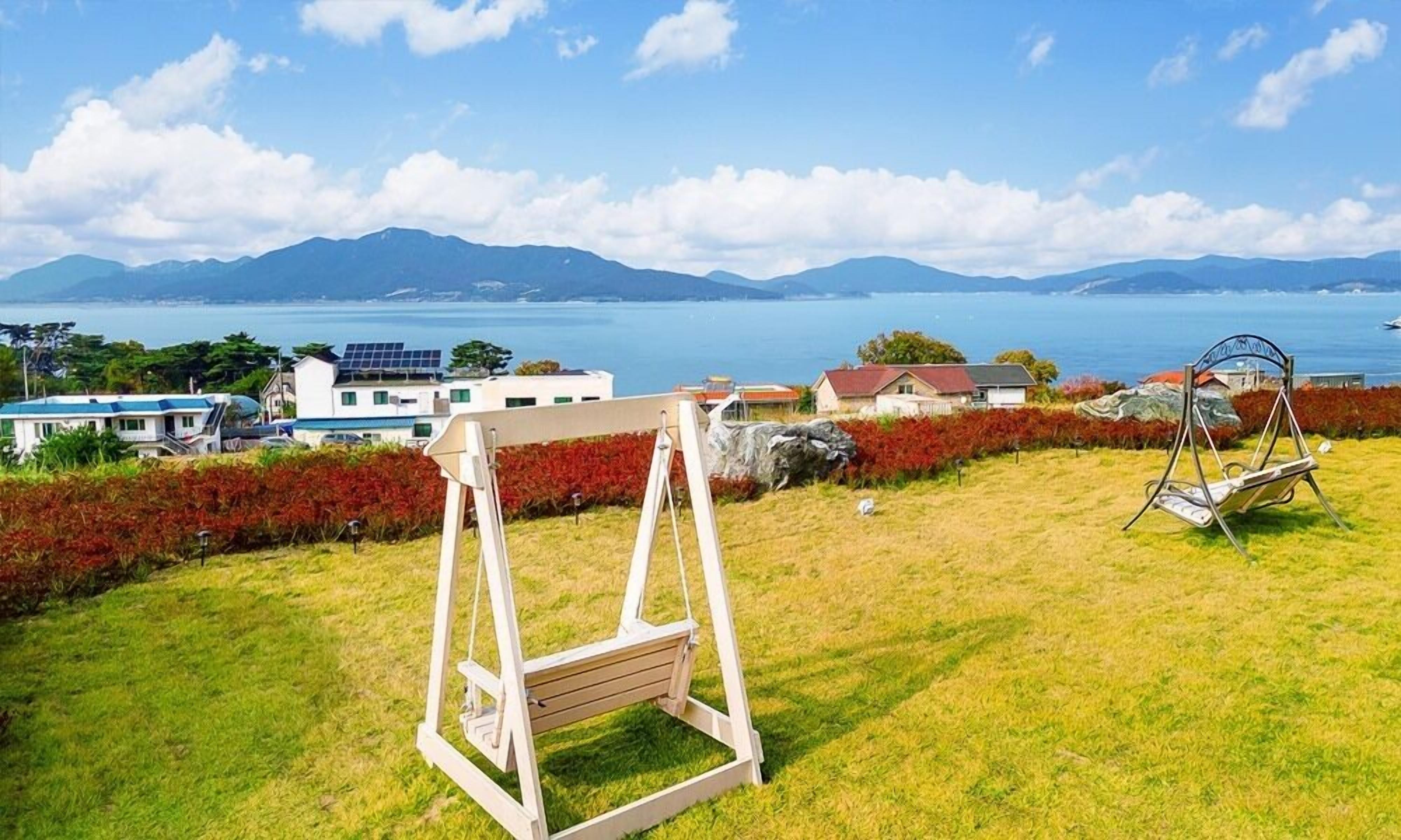 A grassy area with a wooden swing set and a metal sculpture, overlooking a coastal landscape with buildings and mountains.