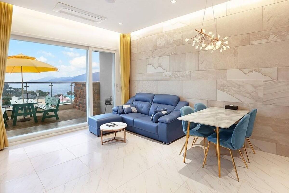 A modern living room with a blue sofa, a round coffee table, and a dining table with chairs. There is a large window with a view of the sea and mountains.