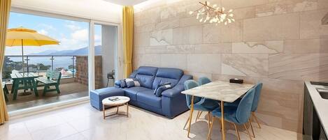 A modern living room with a blue sofa, a round coffee table, and a dining table with chairs. There is a large window with a view of the sea and mountains.