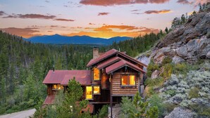 A wooden cabin with a metal roof, surrounded by dense forest and mountains in the background.