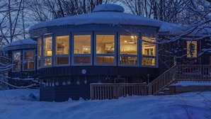 A snow-covered wooden cabin with large windows, a deck, and a staircase leading to the entrance.