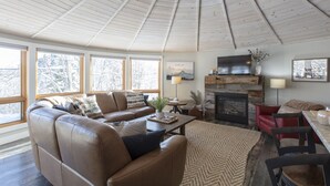 A cozy living room with a fireplace, leather sofas, a coffee table, and large windows offering a view of snow-covered trees.
