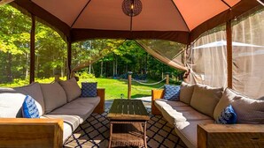 An outdoor seating area with a canopy, cushioned sofas, a wooden table, and a hammock in the background.