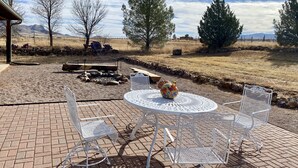 An outdoor patio with a round table and white metal chairs, surrounded by a stone wall and a view of a dry landscape with trees and mountains in the distance. - Sunset Ride Ranch /Foreman's house (Elgin)