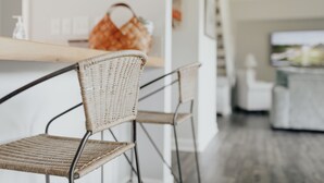 A wicker chair with metal legs, a wooden table, and a wicker basket in a modern kitchen with a white countertop and a view of a living area. - Sunset Villa Across the Street from Beach & Walk to Pool / Golf
 (Johns Island)