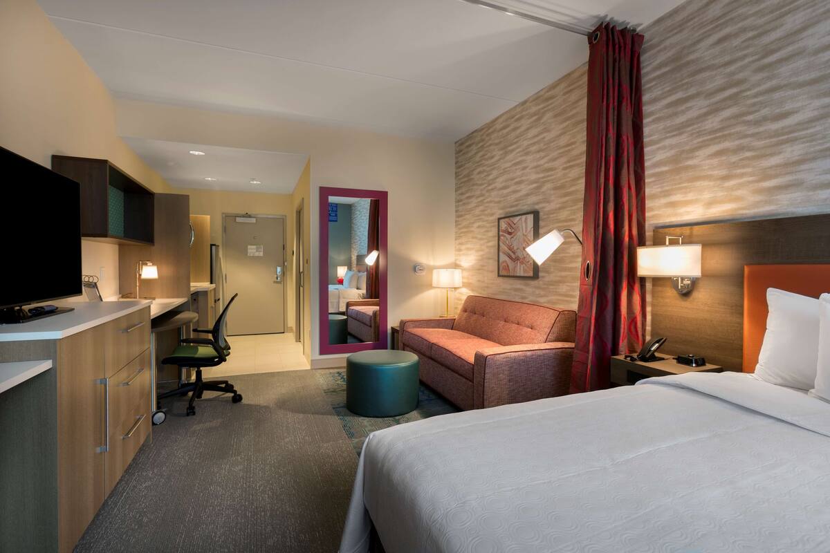 A hotel room with a bed, a desk, a chair, a television, and a bathroom visible through an open door.