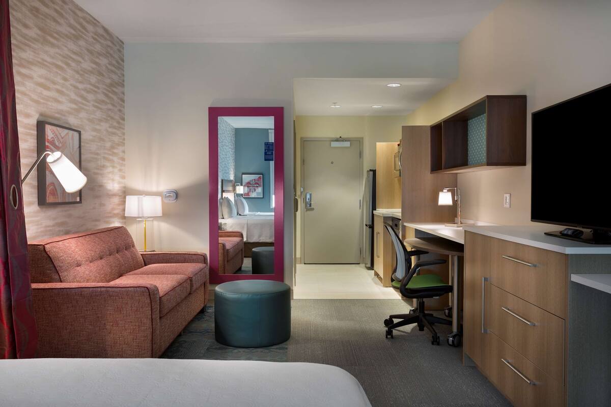A hotel room with a bed, a sofa, a desk with a computer, a TV, and a mirror.