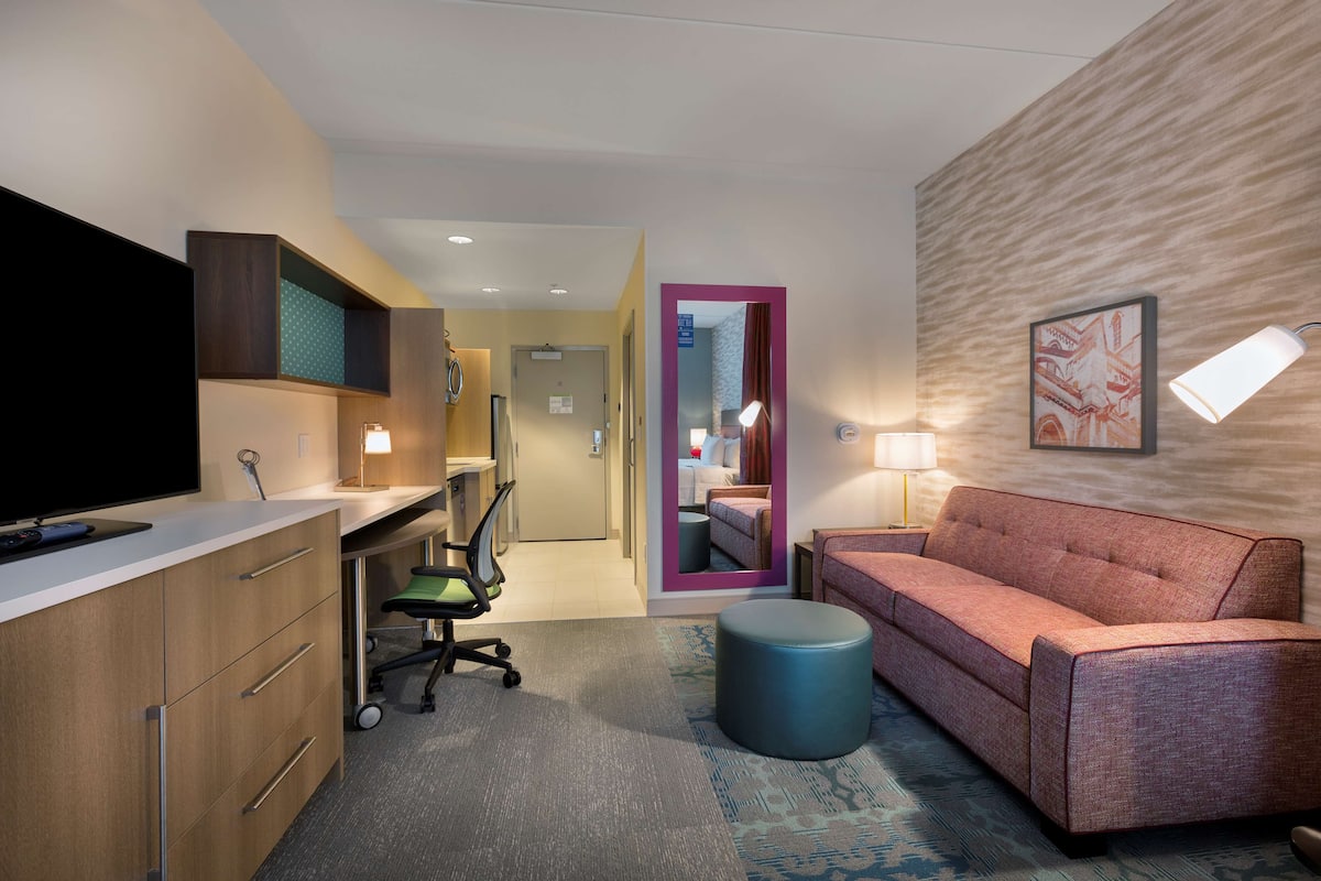 A hotel room with a desk, a sofa, a television, a mirror, and a lamp.