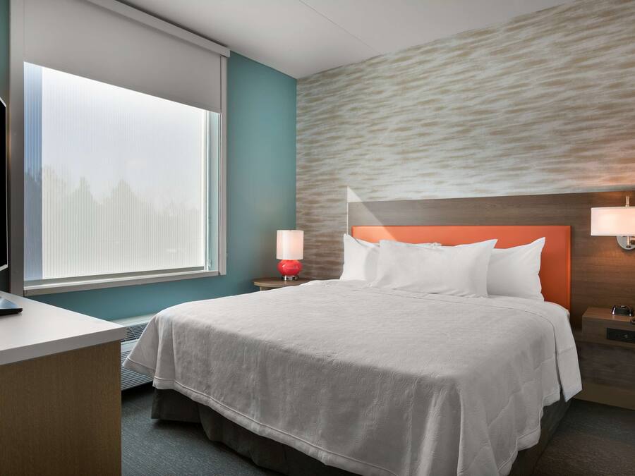 Quarto de hotel com uma cama, uma escrivaninha, uma TV, uma janela com vista para edifícios e uma parede com textura.