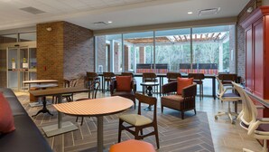 Un espace intérieur vaste et lumineux, aménagé avec différents types de sièges, notamment des tables et des chaises, de grandes fenêtres laissant entrer la lumière naturelle et un mur en briques.