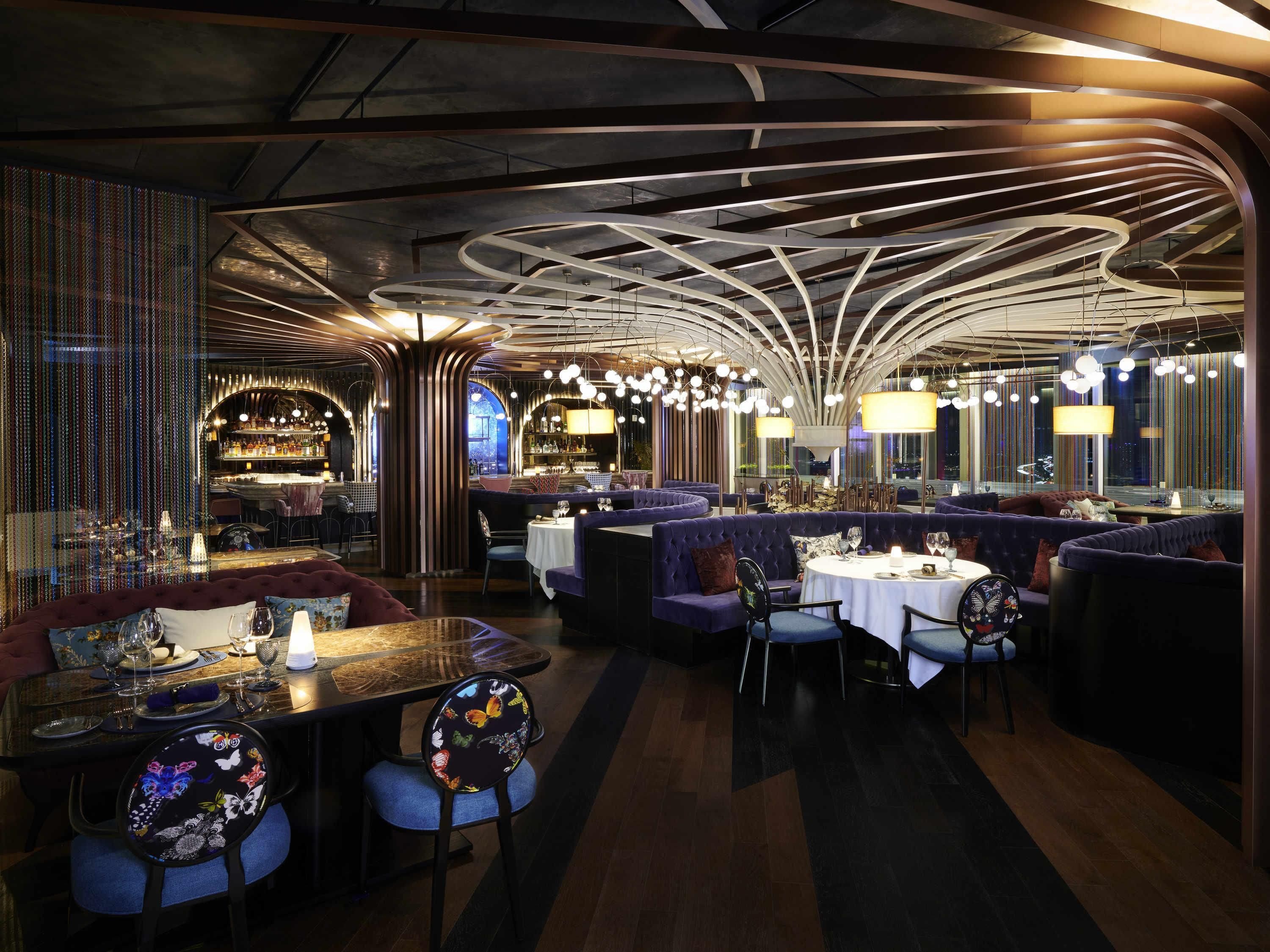 A restaurant interior with round tables, cushioned seating, and a modern ceiling design.