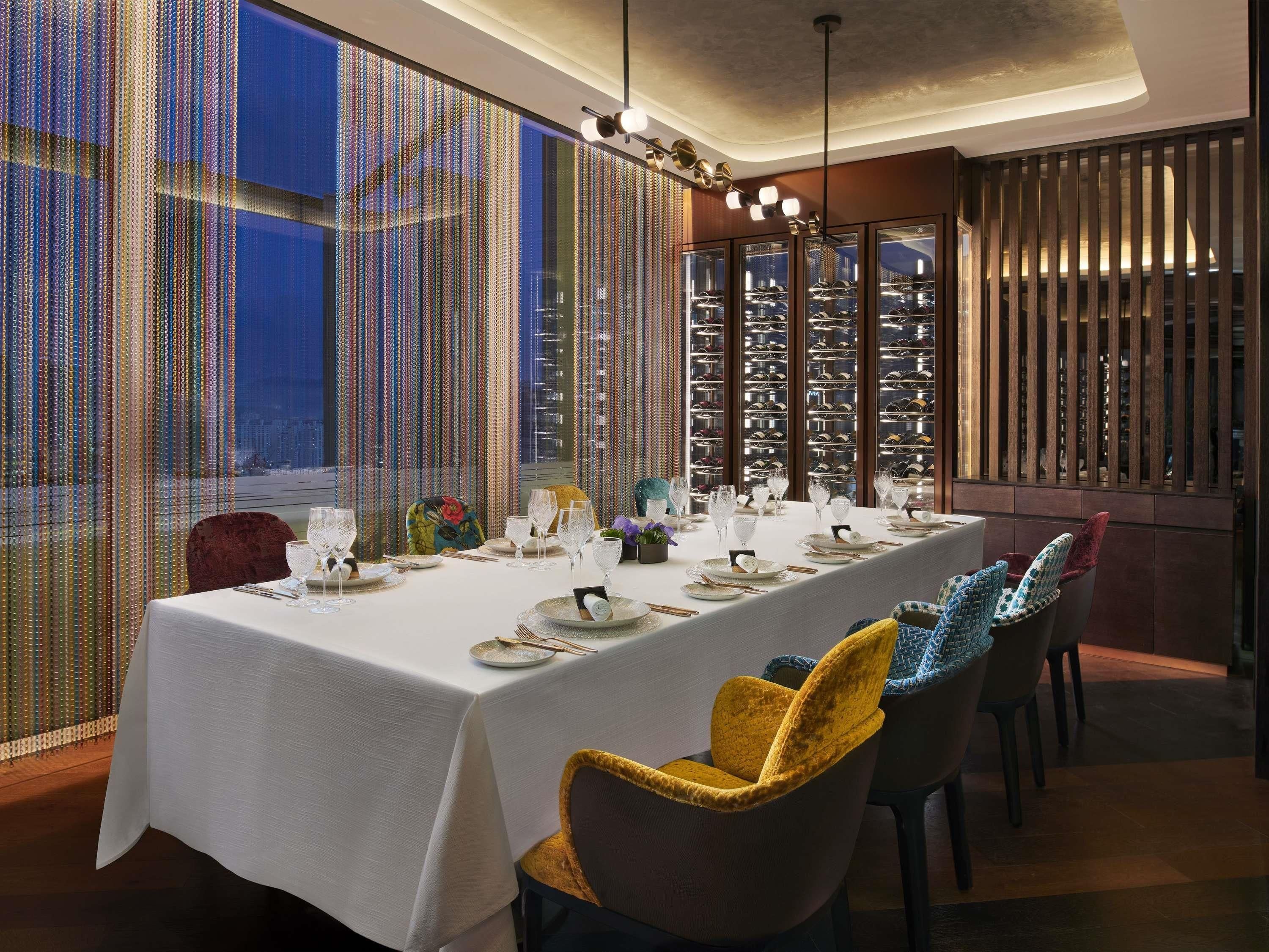 A dining area with a long table set for a meal, chairs with patterned cushions, and a view of a cityscape through a patterned window.