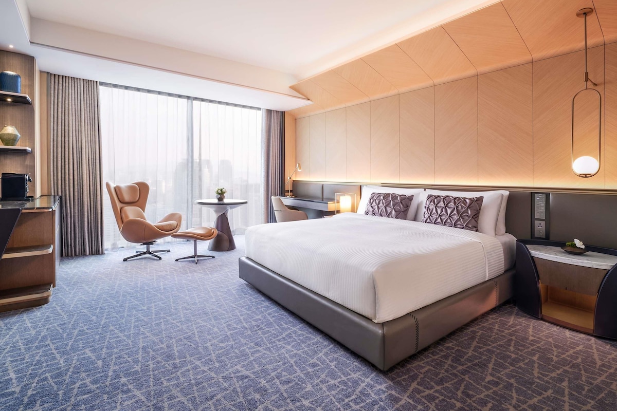 Une chambre d’hôtel moderne dotée d’un grand lit, d’un bureau, d’une chaise et offrant une vue sur la ville grâce à de grandes fenêtres.