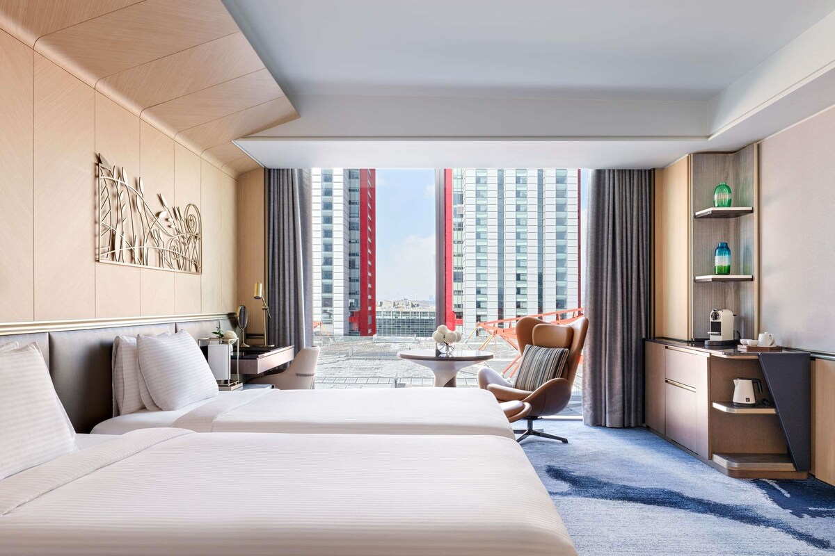 Une chambre d’hôtel avec deux lits, un bureau, une chaise, une petite table et une vue sur le paysage urbain.