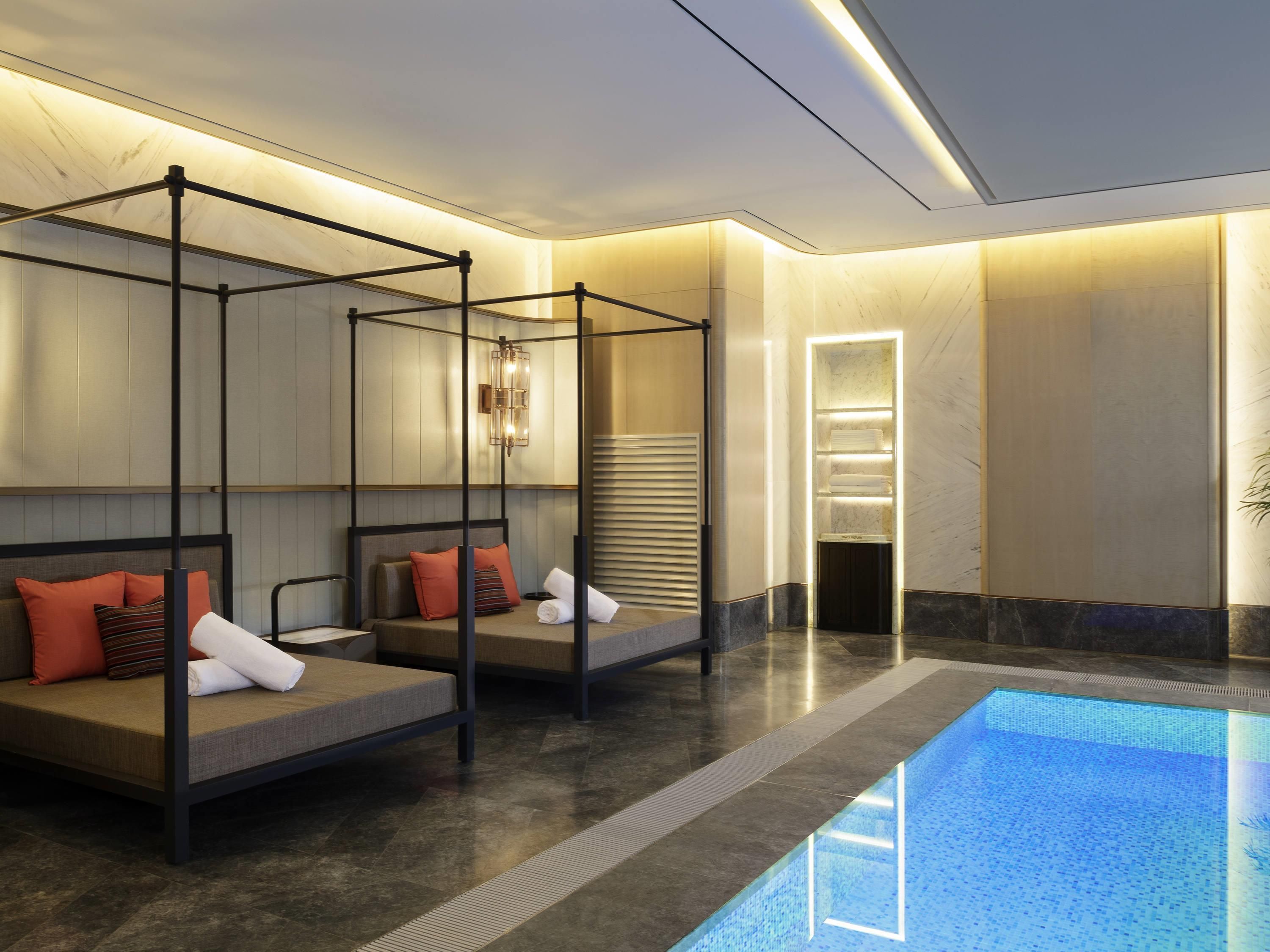 A modern hotel room with a pool, two beds with headboards, a vanity with a mirror, and a ceiling light fixture.