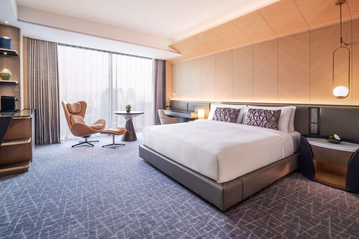 Une chambre d’hôtel moderne dotée d’un grand lit, d’un bureau, d’une chaise et offrant une vue sur la ville grâce à de grandes fenêtres.
