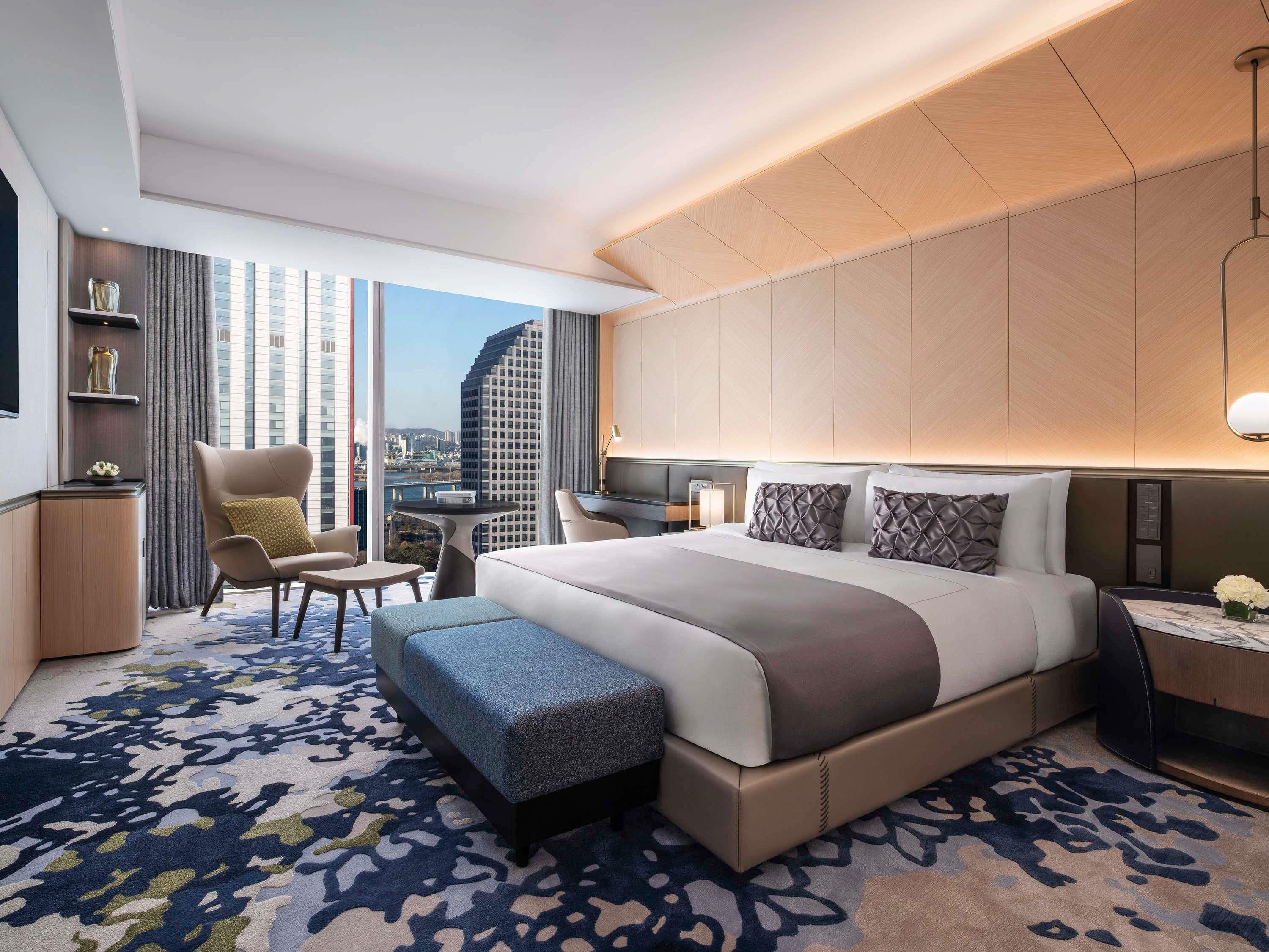 A modern hotel room with a large bed, a desk, a chair, and a view of the cityscape.