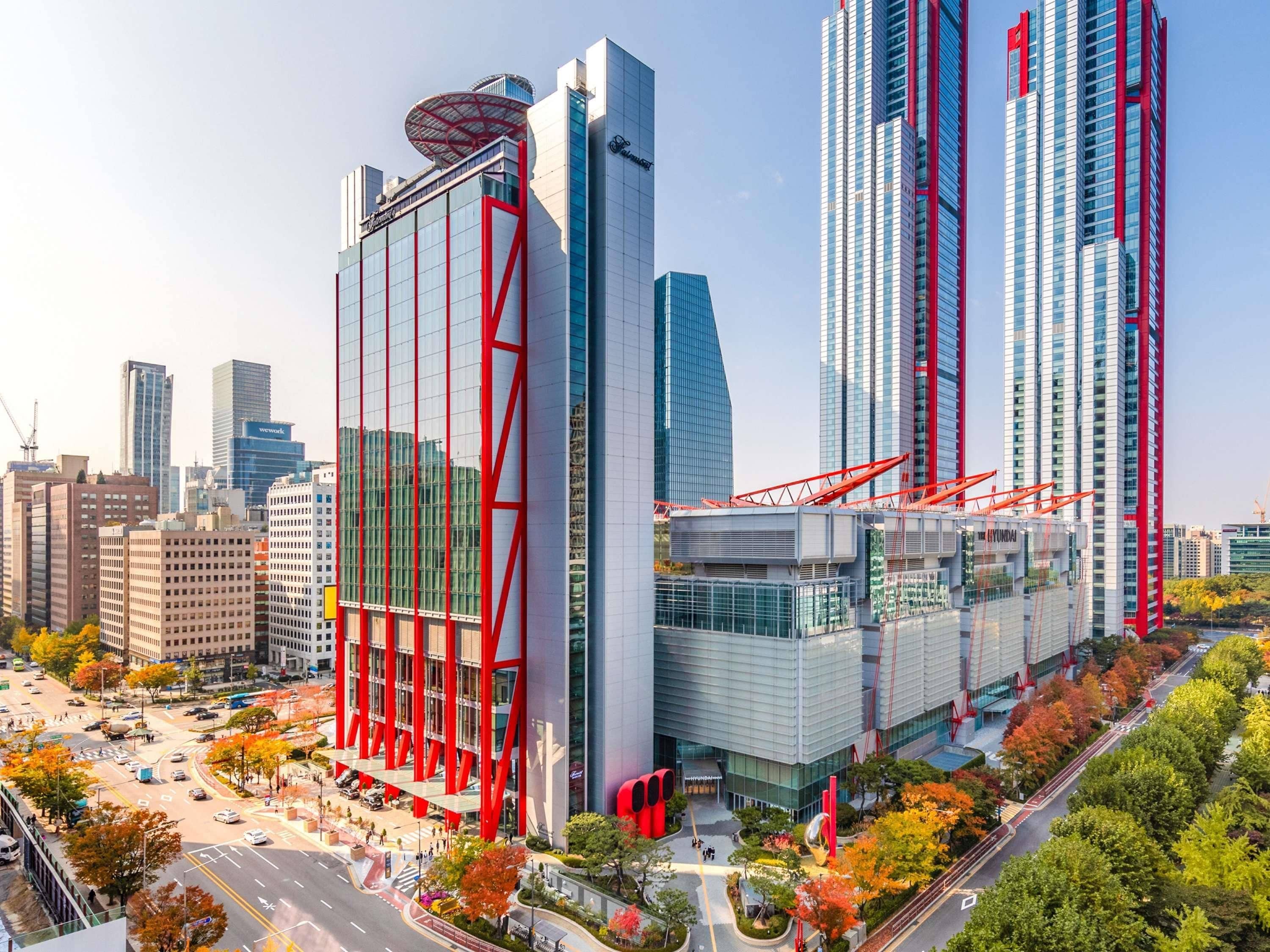 A modern high-rise building with a distinctive red structural design, surrounded by other tall buildings and a busy urban street.