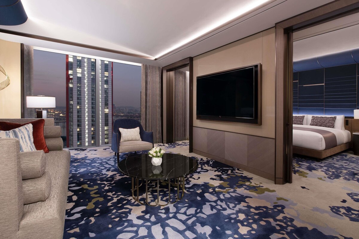 A modern hotel room with a large window, a flat-screen TV, a dining table, and a sofa.