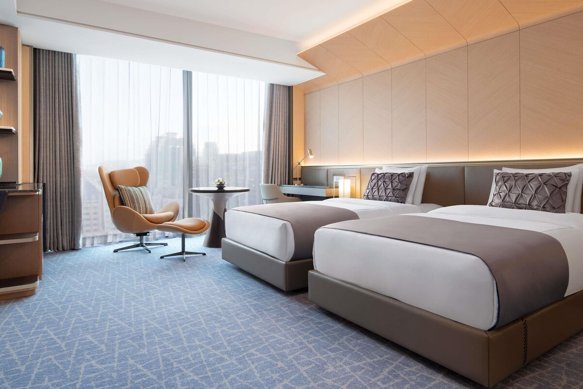 Une chambre d’hôtel avec deux lits, un bureau, une chaise et une grande fenêtre avec des rideaux.