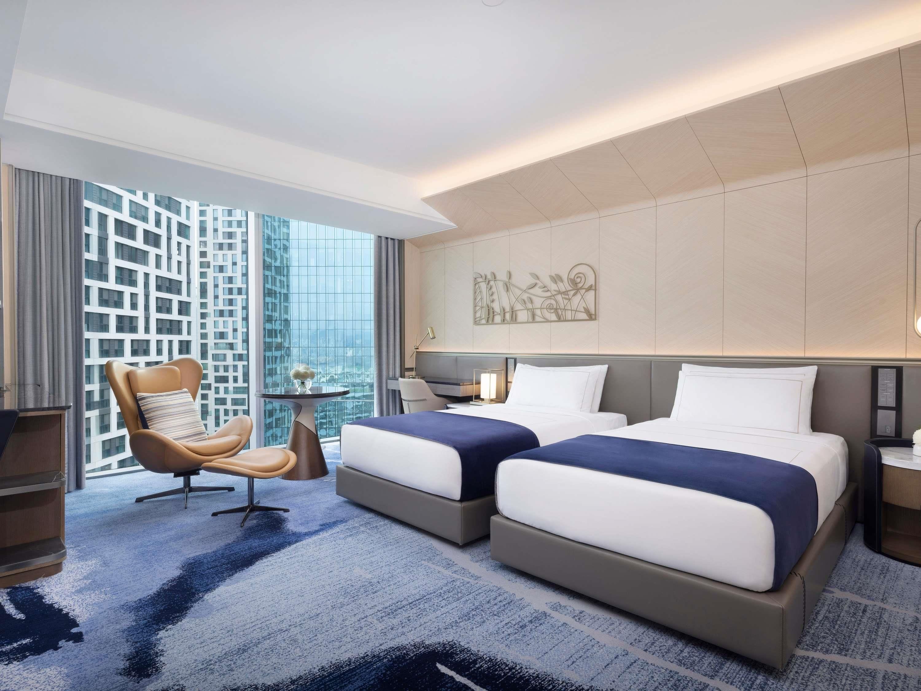 A hotel room with two beds, a chair, a small table, and a view of a cityscape.