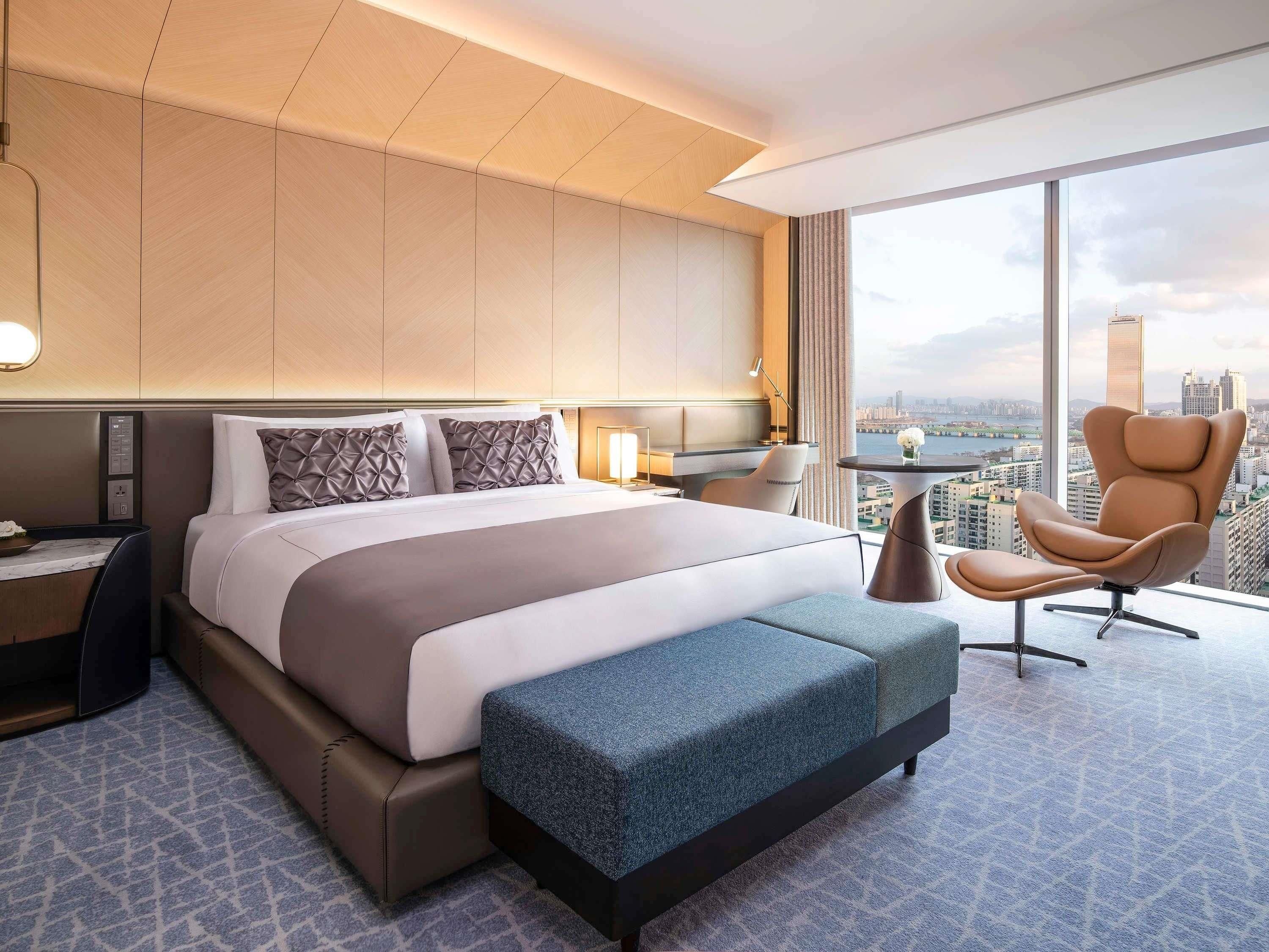 Ett modernt hotellrum med en stor säng, en sittgrupp med en fåtölj och ett litet bord, ett skrivbord och ett stort fönster med utsikt över staden.