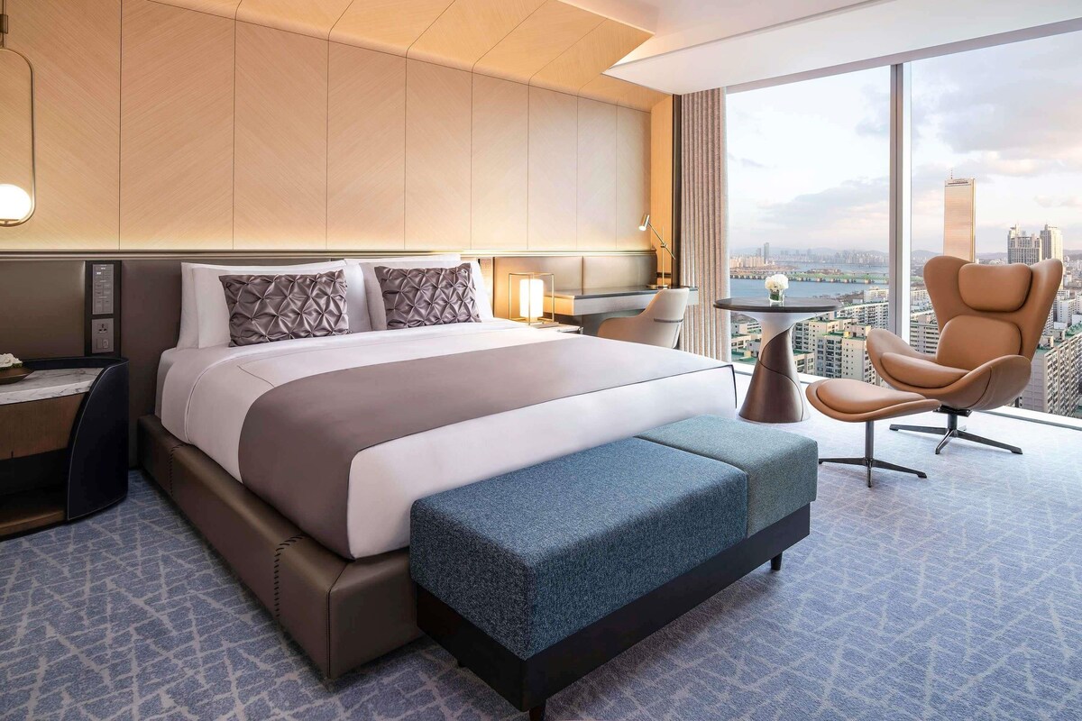 Une chambre d’hôtel moderne dotée d’un grand lit, d’un coin salon avec un fauteuil et un pouf, d’un bureau et d’une grande fenêtre donnant sur la ville.