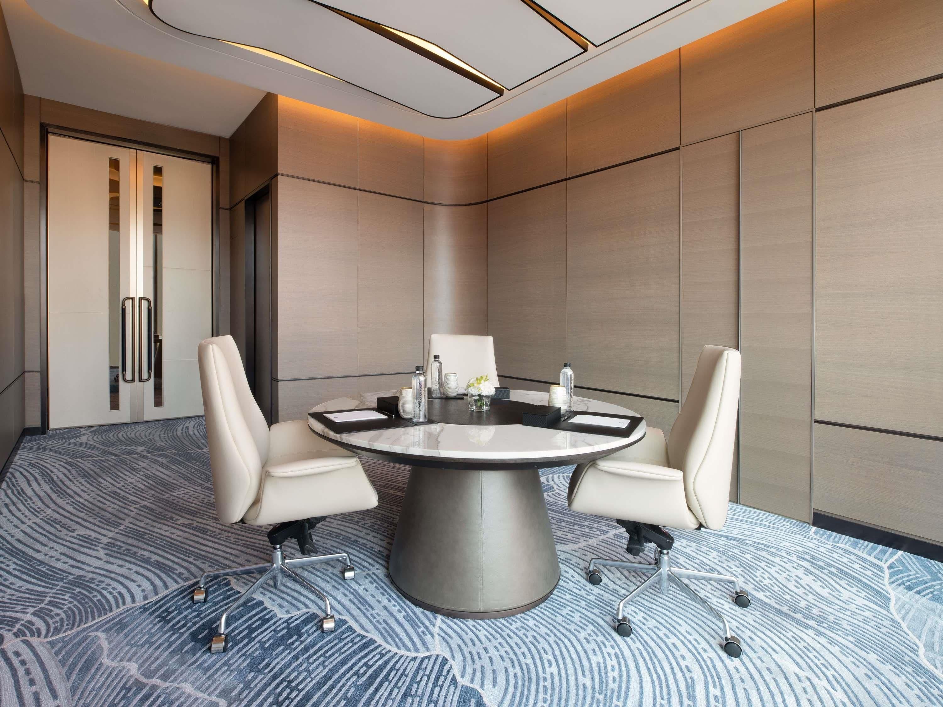 A conference room with a round table, four chairs, a glass partition, and a patterned carpet.