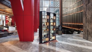 Een ruime lobby met een modern design, met een grote glazen wand, een rood structureel element en een goed verlicht gebied met een boekenkast.