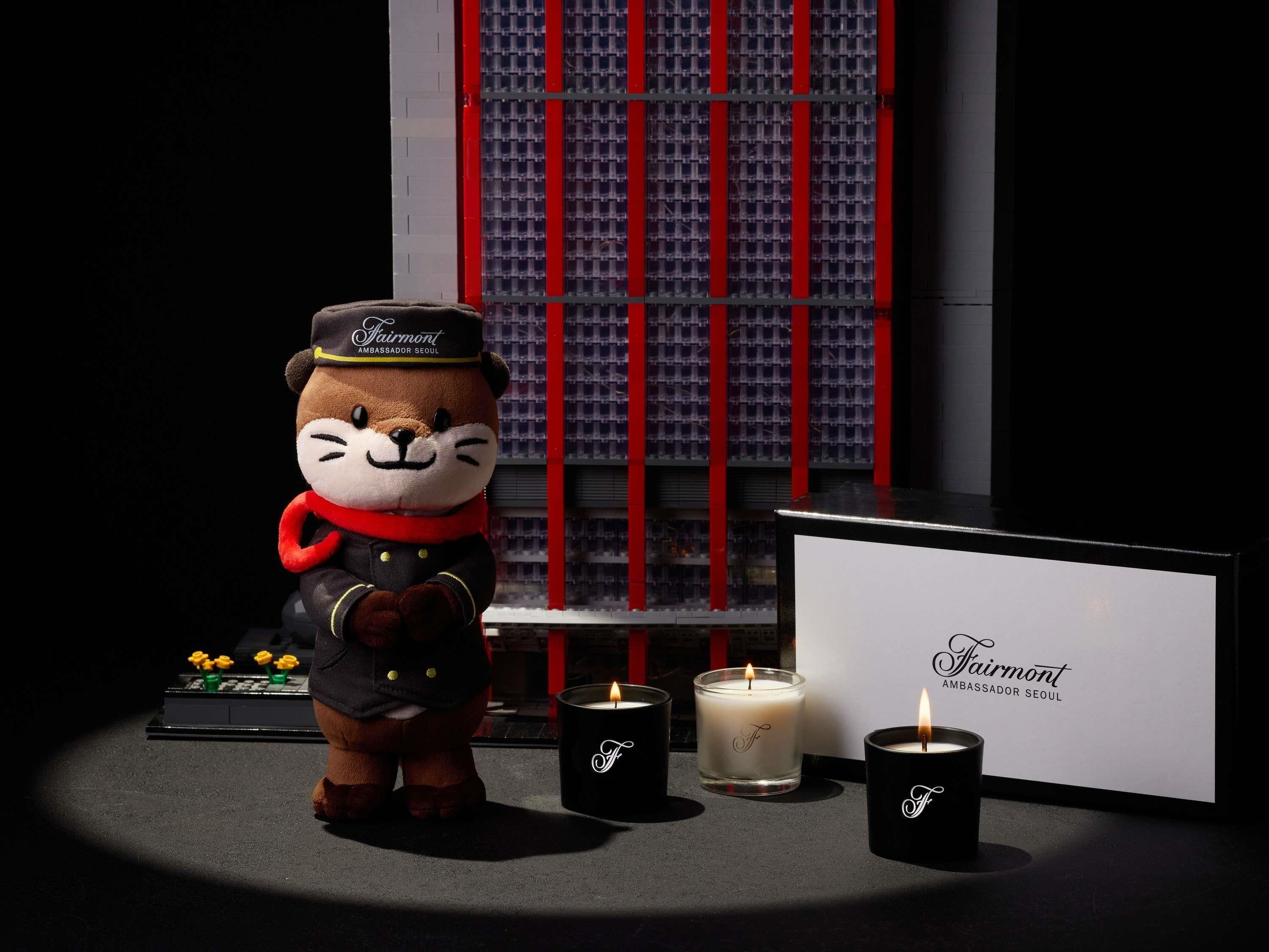 A plush toy bear with a cap and scarf, three candles with 'Fairmont' branding, and a backdrop of a red and white patterned structure.