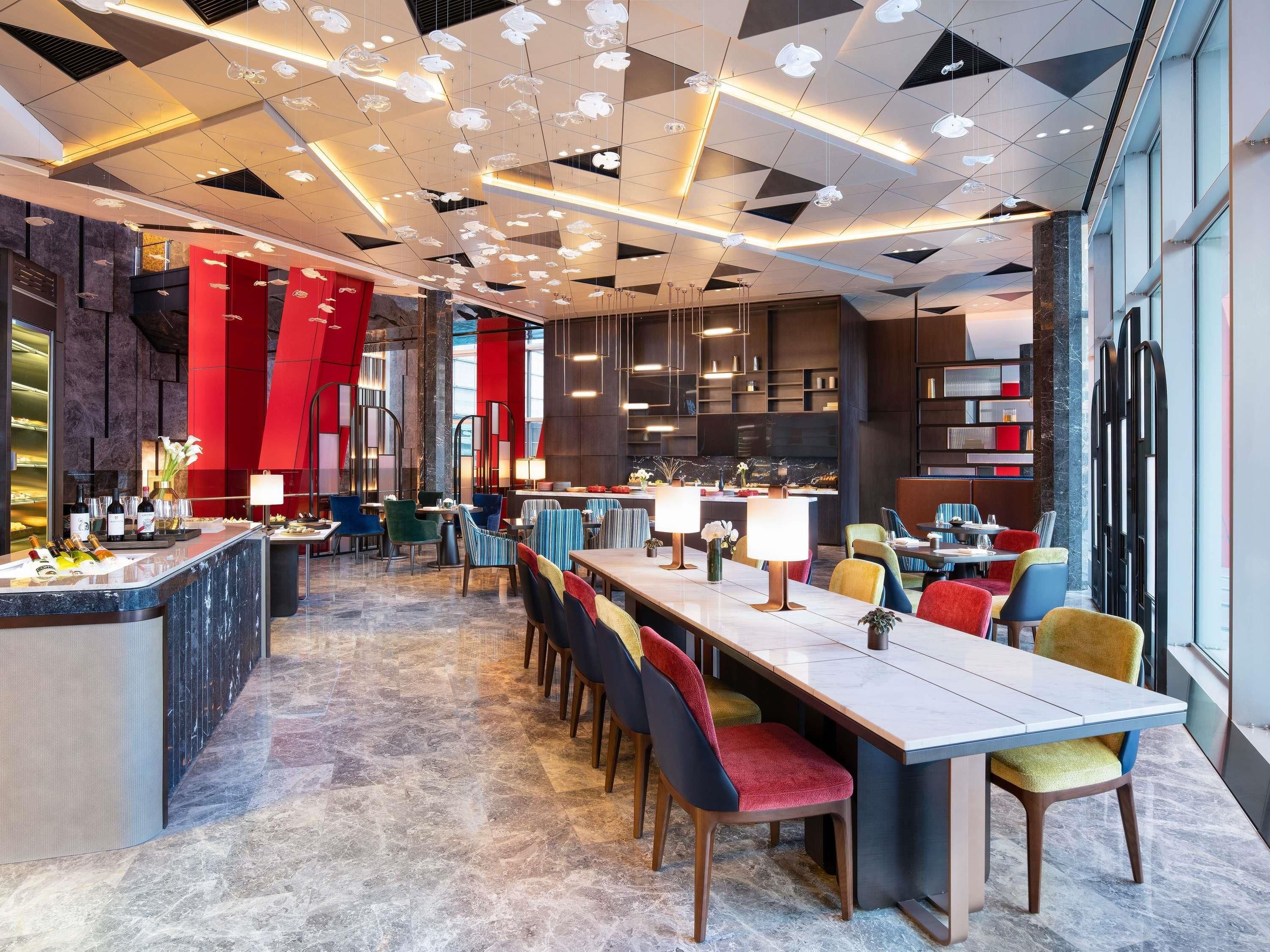 A modern restaurant with a long communal table, colorful chairs, and a bar area with bottles and glasses.