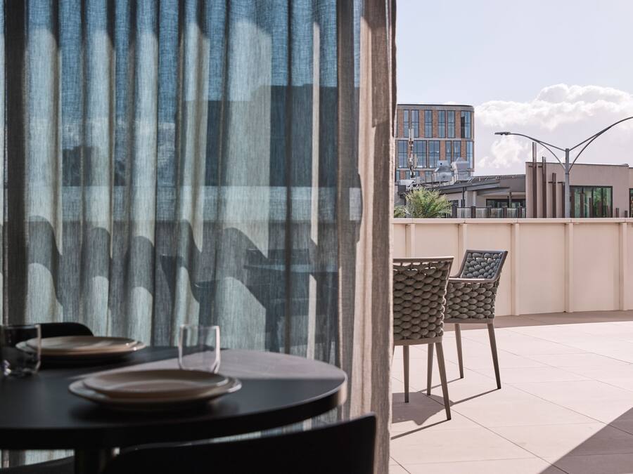 Un coin repas avec une table dressée pour deux personnes, un verre d’eau et une assiette. Il y a deux chaises au design tressé. Dehors, on aperçoit un bâtiment au design moderne et un ciel dégagé avec quelques nuages.