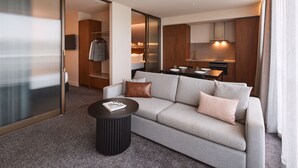 Una habitación de hotel moderna con un sofá gris, una mesa de centro negra redonda y una cocina visible a través de una puerta abierta.