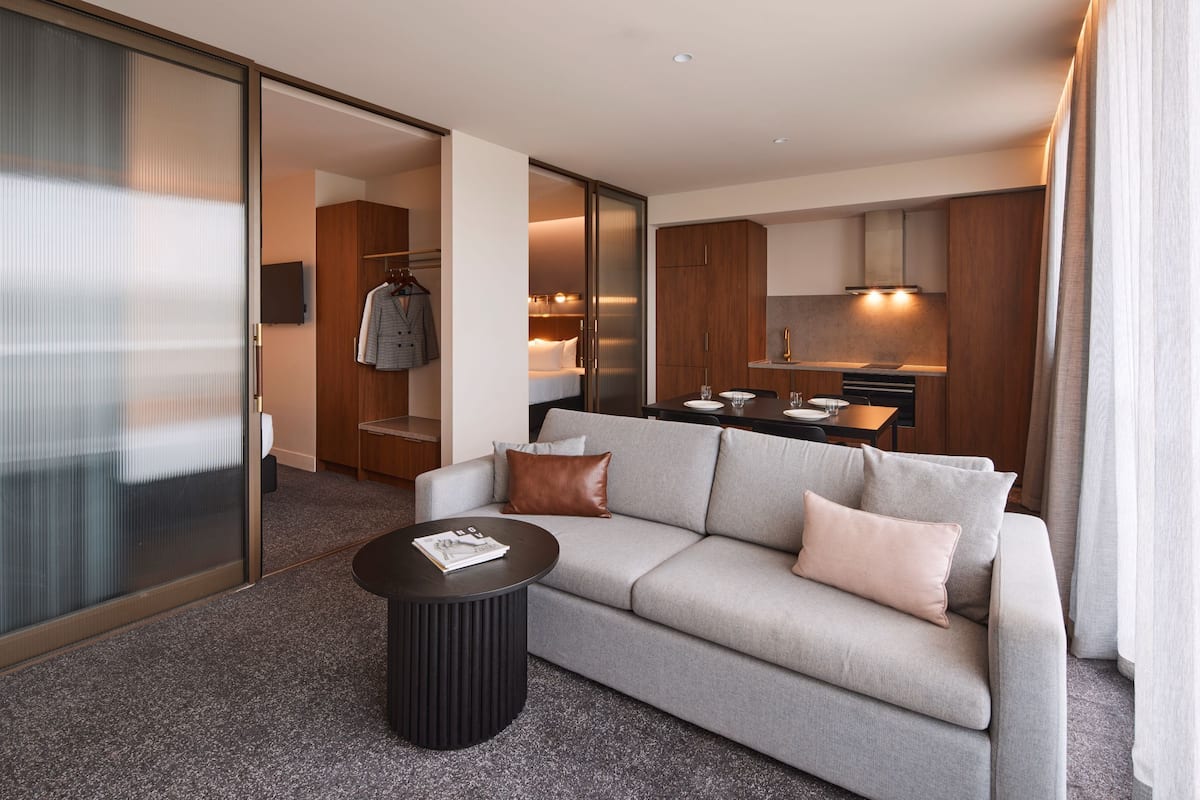 Et moderne hotelværelse med en grå sofa, et rundt sort sofabord og et tekøkken, der kan anes gennem en åben dør.