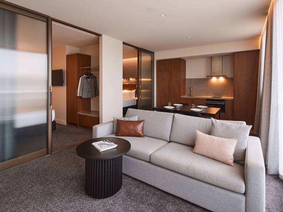 A modern hotel room with a grey sofa, a round black coffee table, and a kitchen area visible through an open door.