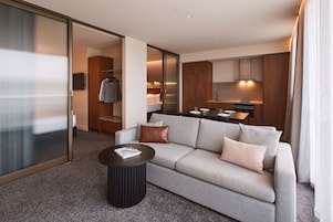 Une chambre d’hôtel moderne avec un canapé gris, une table basse ronde noire et un coin cuisine visible à travers une porte ouverte.