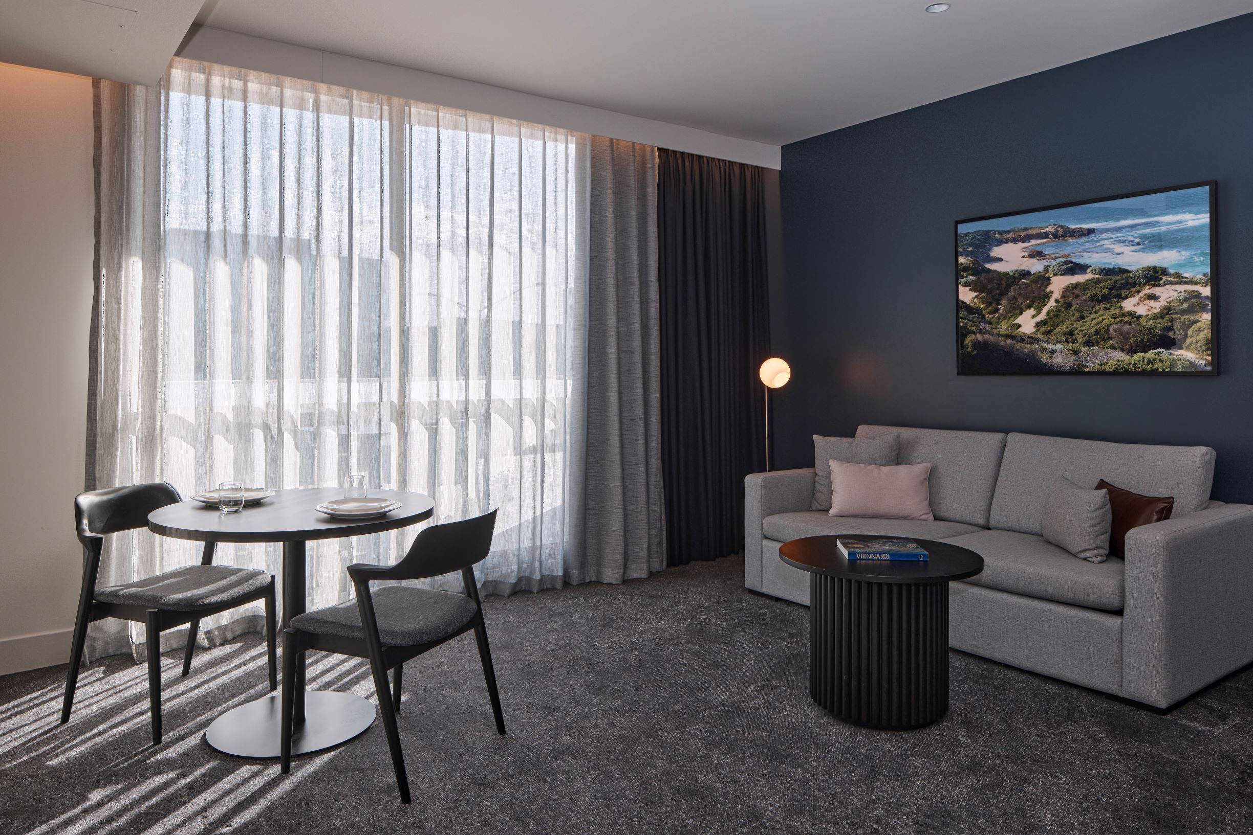 Una habitación de hotel moderna con una mesa de comedor redonda, dos sillas, un sofá gris, una mesa de centro y un ventanal con cortinas translúcidas.
