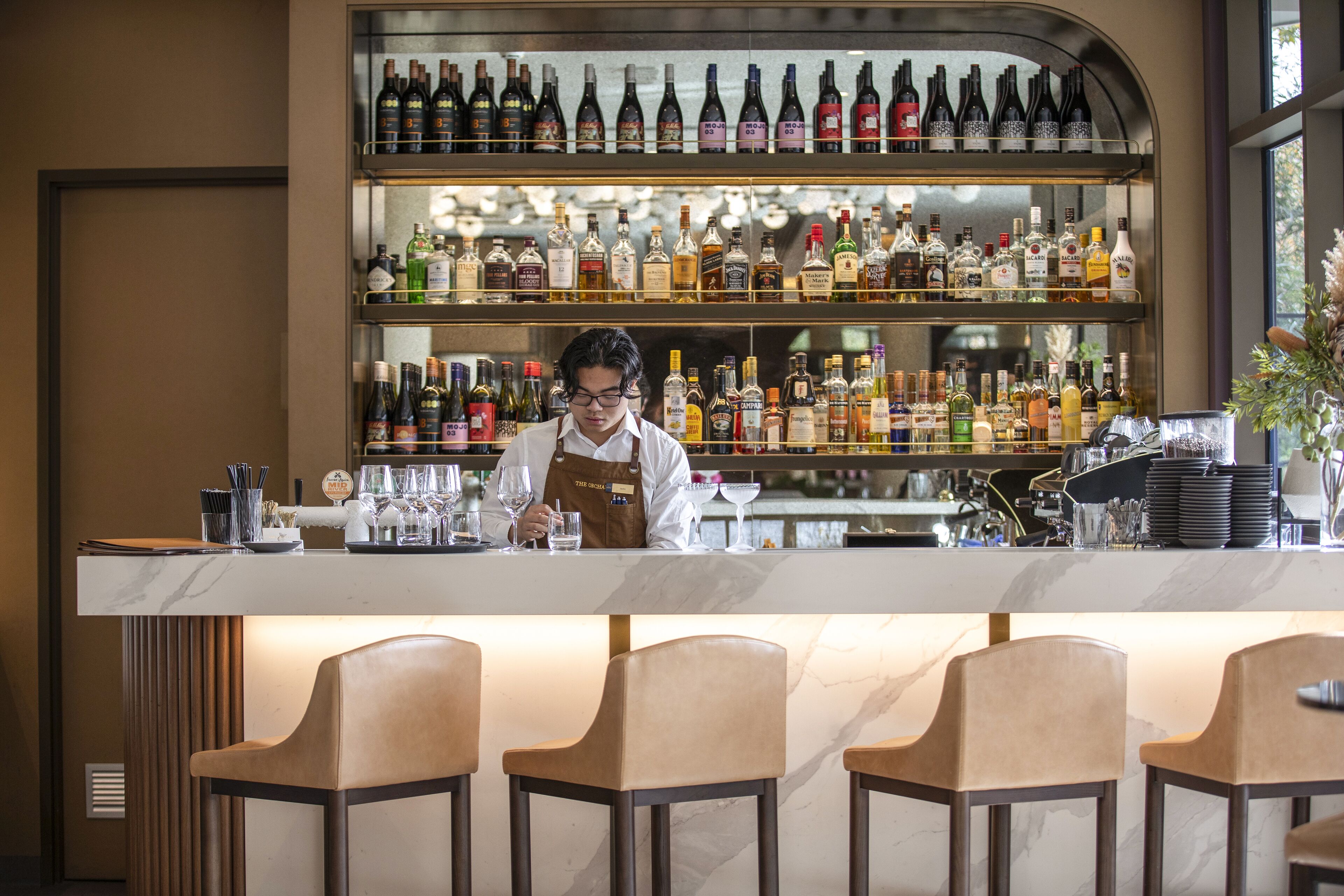 A bar area with a bartender, shelves of various liquor bottles, and bar stools.