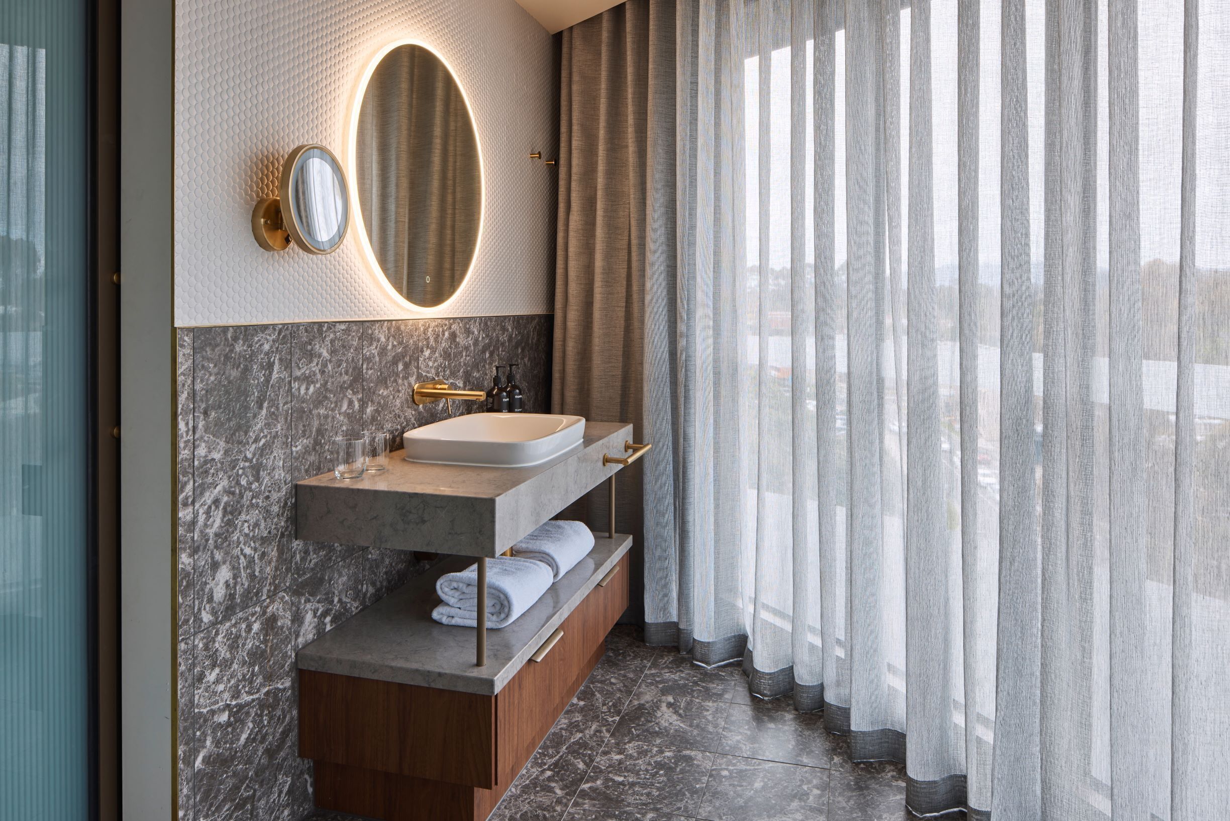 Une salle de bain moderne avec un meuble-lavabo en marbre, un grand miroir et une vue à travers des rideaux transparents.