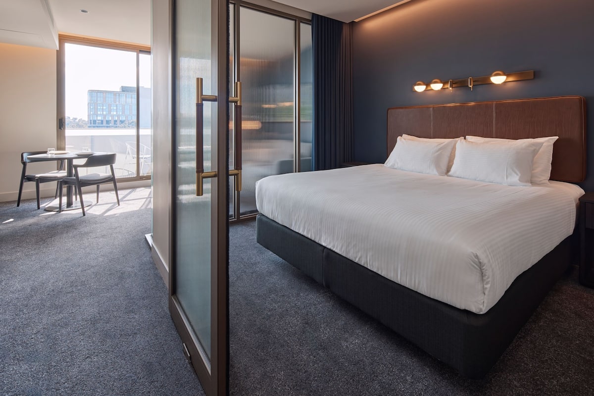 Et hotelværelse med en stor seng, et skrivebord med stol, en skydedør og udsigt over byen.