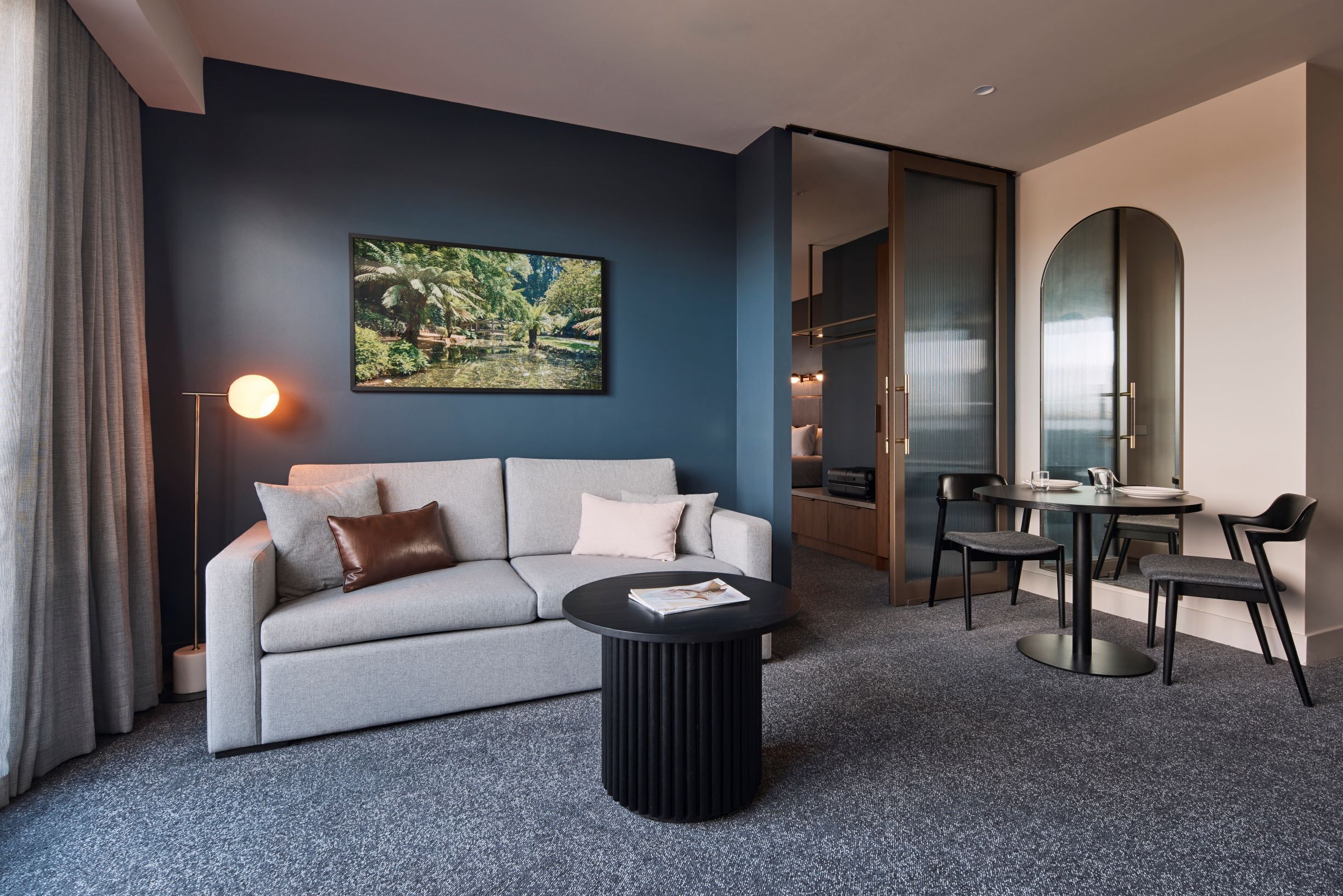 A modern hotel room with a grey sofa, a round black coffee table, and a dining table with chairs. A large framed picture of a tropical scene hangs on the wall.