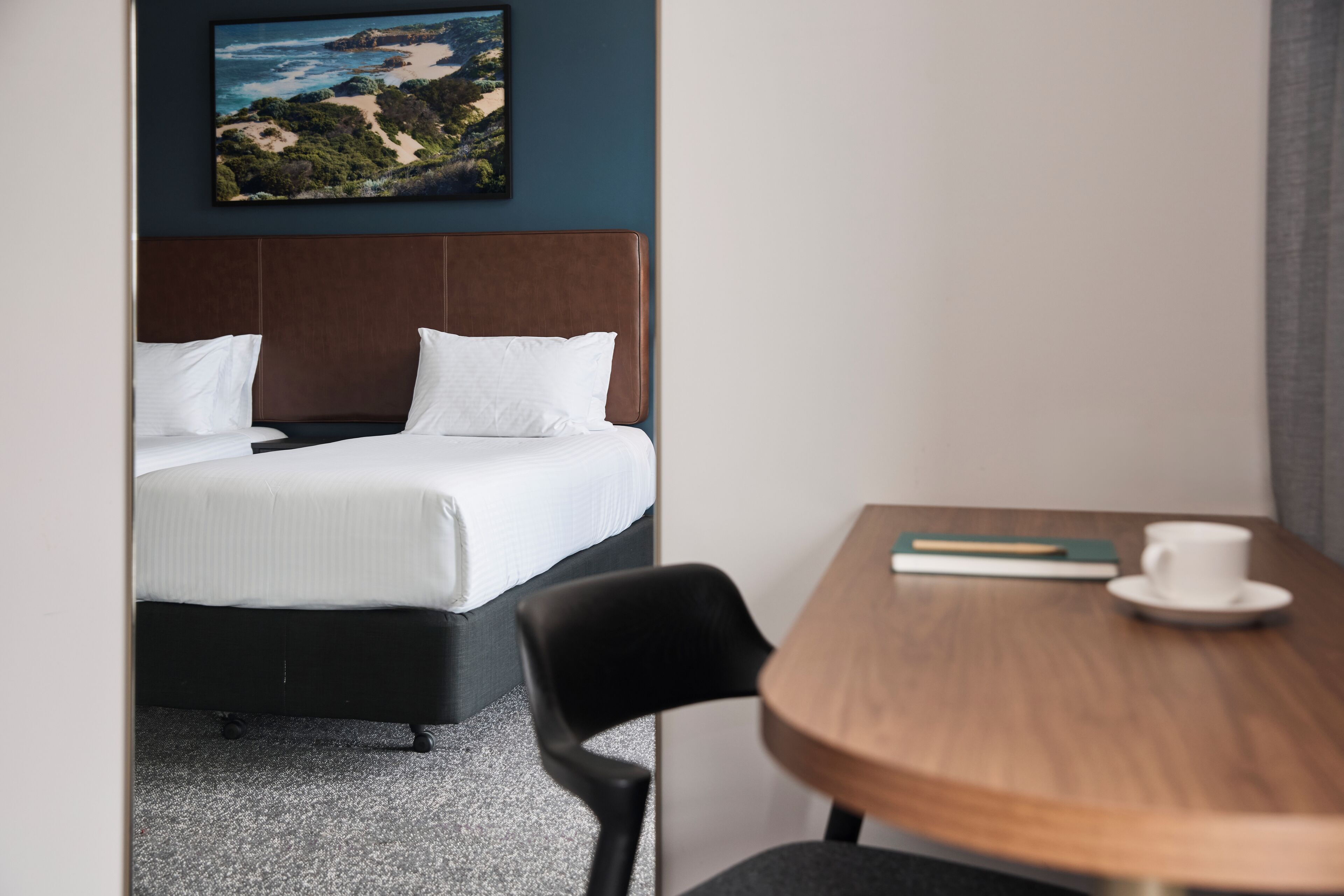 Ein Hotelzimmer mit einem Bett, einem Holztisch mit Tasse und Buch sowie einem Strandbild an der Wand.
