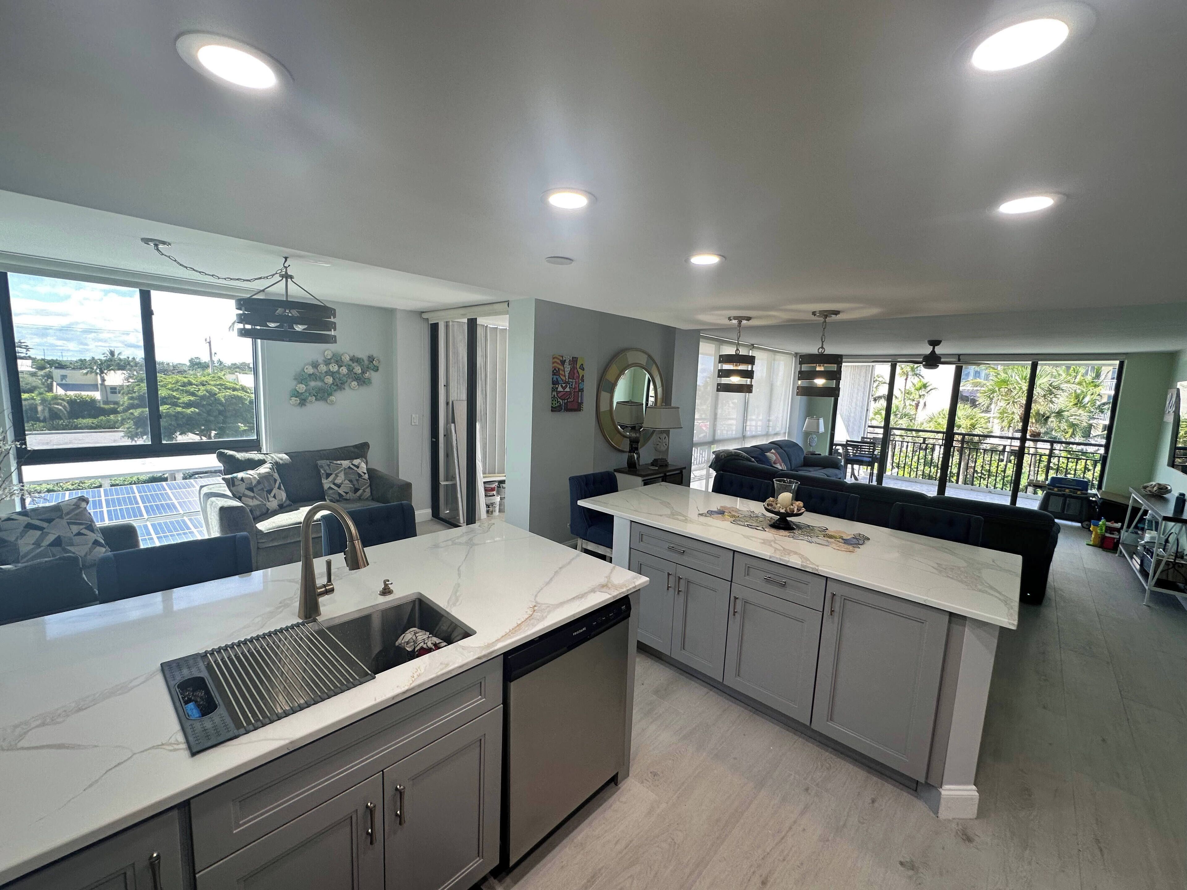 A modern kitchen with a marble countertop, grey cabinets, and a built-in dishwasher. There is a dining area with a blue sofa and a dining table. Large windows provide a view of the outdoors.