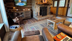 A cozy living room with a stone fireplace, a television mounted on the wall, and a comfortable seating area with a rug and leather chairs. - WINDSONG! Peaceful, Quiet Cabin Bordering Nat. Park Near Downtown Gatlinburg (Gatlinburg)