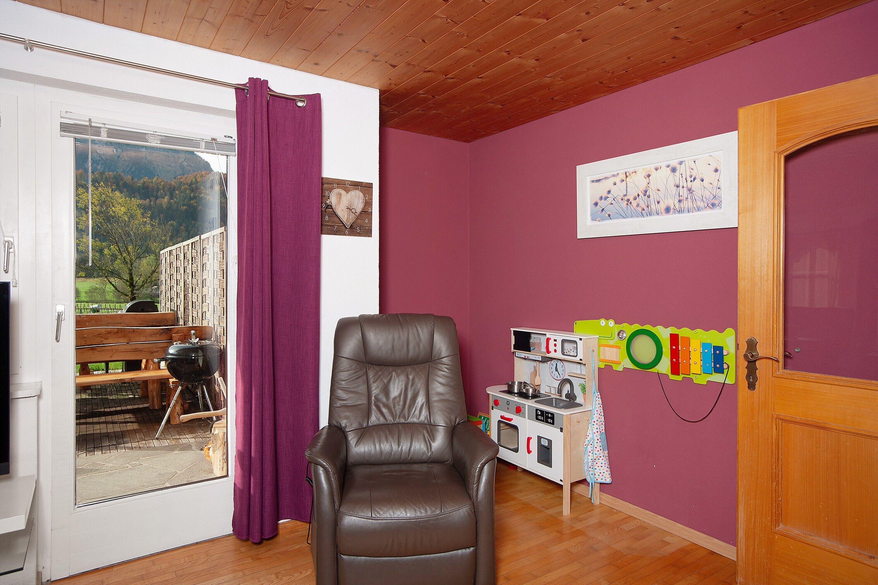 Ein Zimmer mit einer Schiebeglastür, die zu einem Außenbereich führt, einer Holzbalkendecke, einem braunen Ledersessel, einer Spielküche für Kinder und einer Tür mit Holzrahmen.