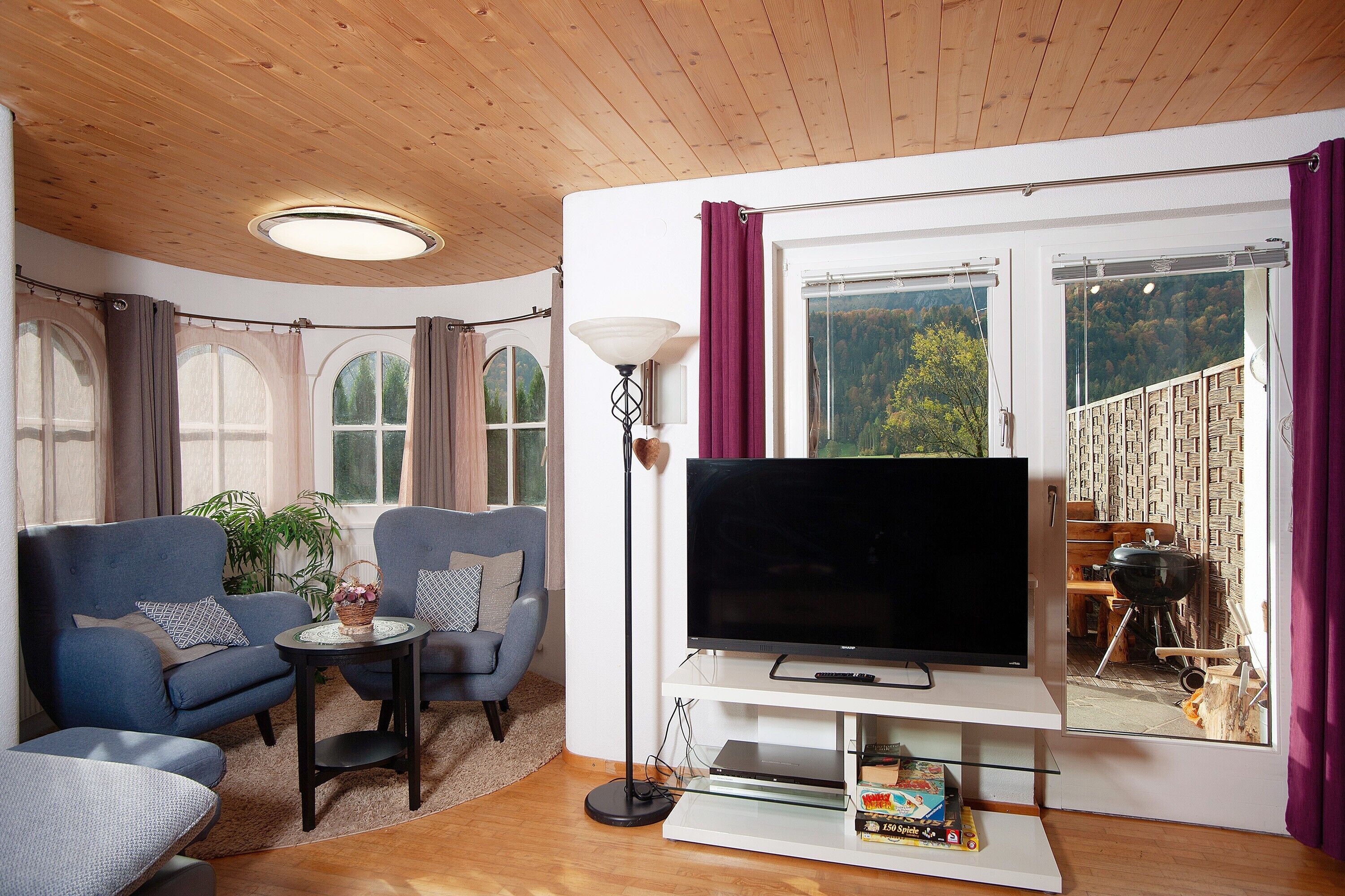 A living room with a TV, sofas, a small table, a lamp, and a large window with curtains.