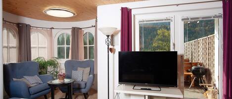 A living room with a TV, sofas, a small table, a lamp, and a large window with curtains.