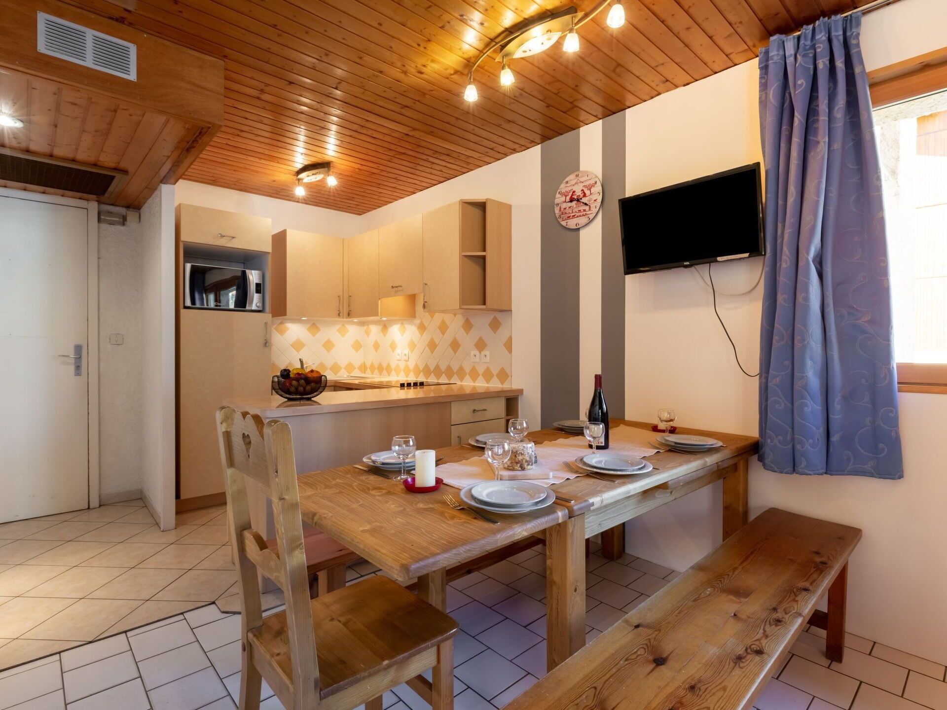 A cozy dining area with a wooden table set for four, a clock on the wall, and a television mounted above the table.