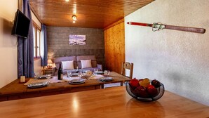 Un coin repas avec une table en bois dressée pour deux personnes, un saladier de fruits et une paire de skis accrochés au mur.